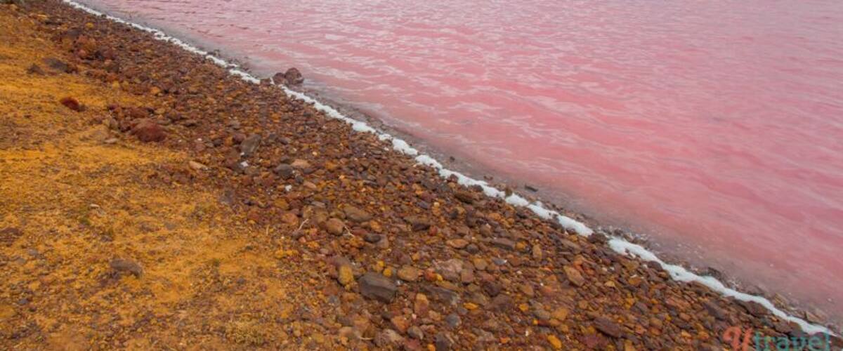 A beach with pink water, pebbles, and a hilly landscape.