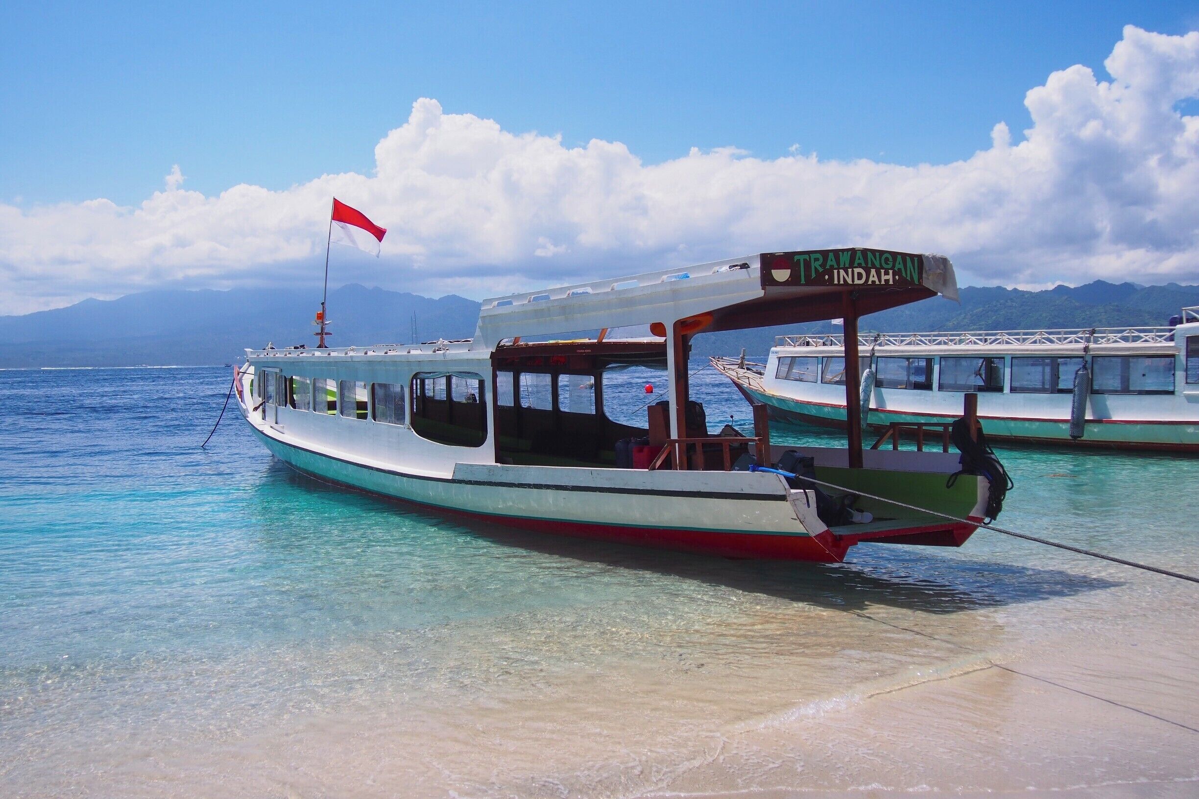 A boat named "TRAWANOAN INDAH" docked at a beach with clear turquoise water and mountains in the background.