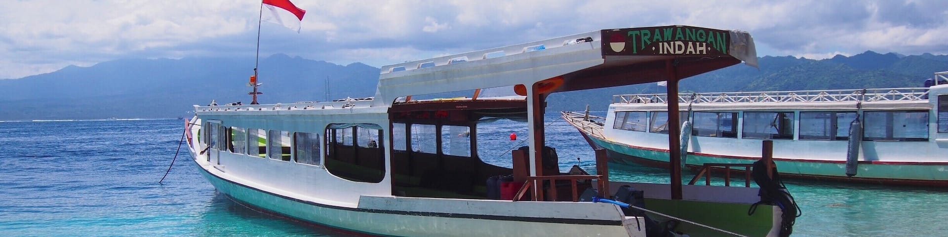 A boat named "TRAWANOAN INDAH" docked at a beach with clear turquoise water and mountains in the background.