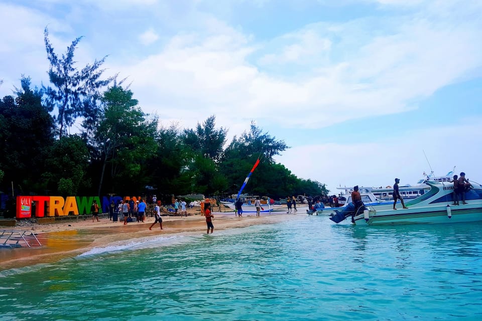 Una playa con agua turquesa cristalina, gente caminando por la orilla y barcos amarrados cerca.