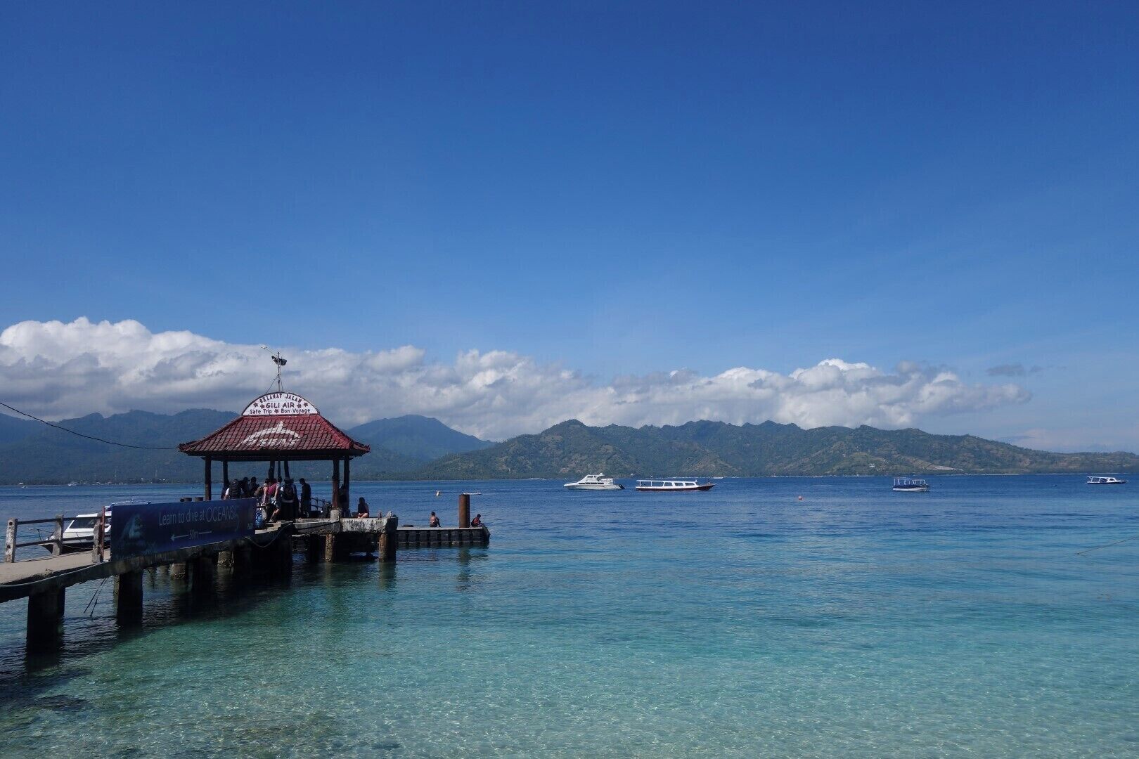 A pier with a covered seating area, clear turquoise water, and boats in the distance with mountains in the background.