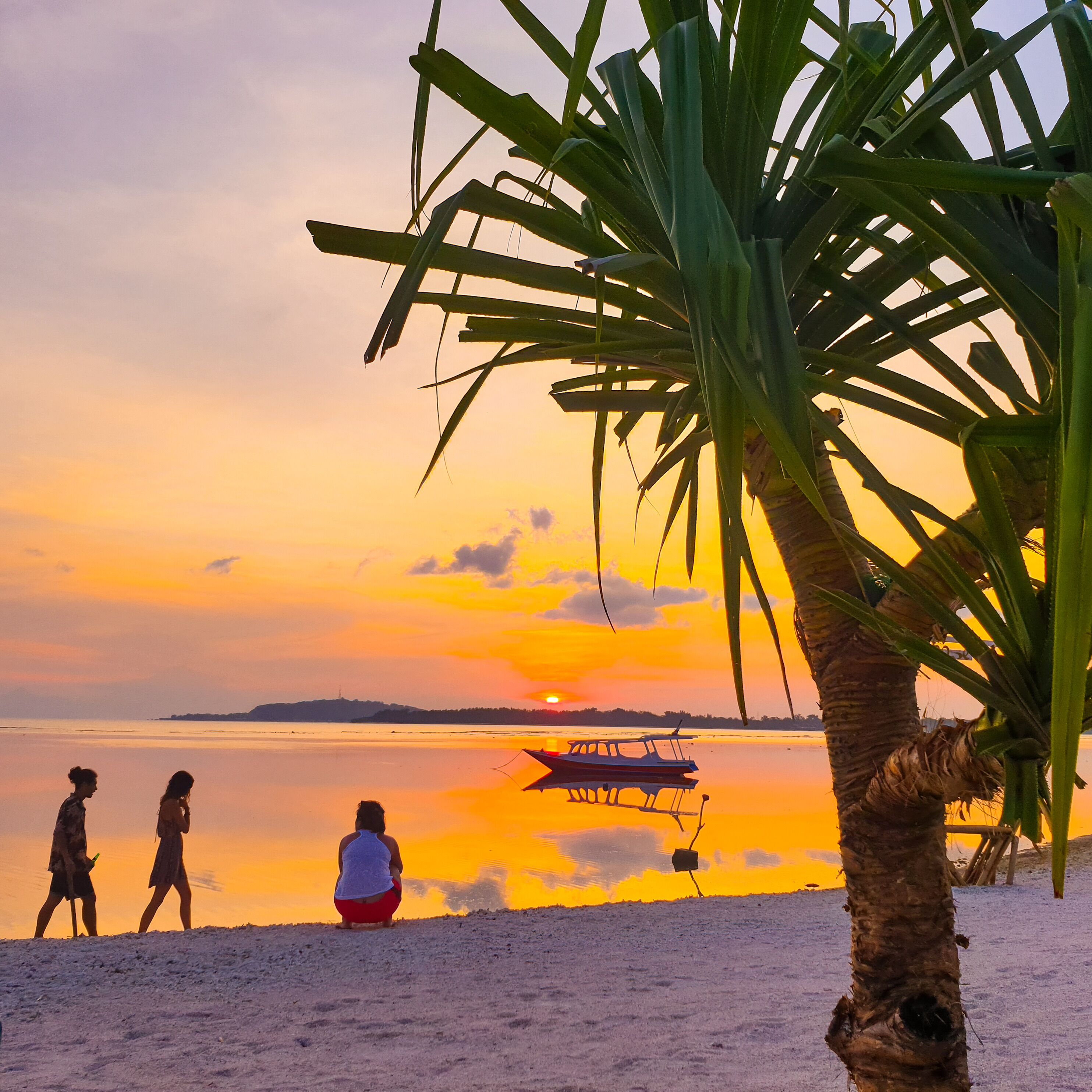 A sunset over a calm beach with people walking and a boat anchored in the water.