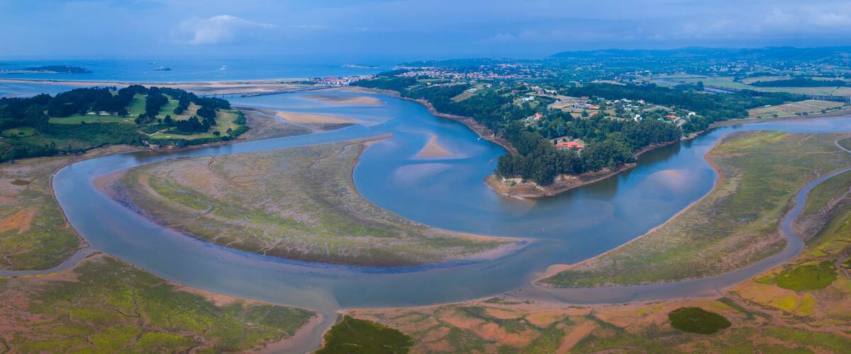 An aerial view of a river delta with multiple channels, surrounded by green landscapes and a small cluster of buildings.