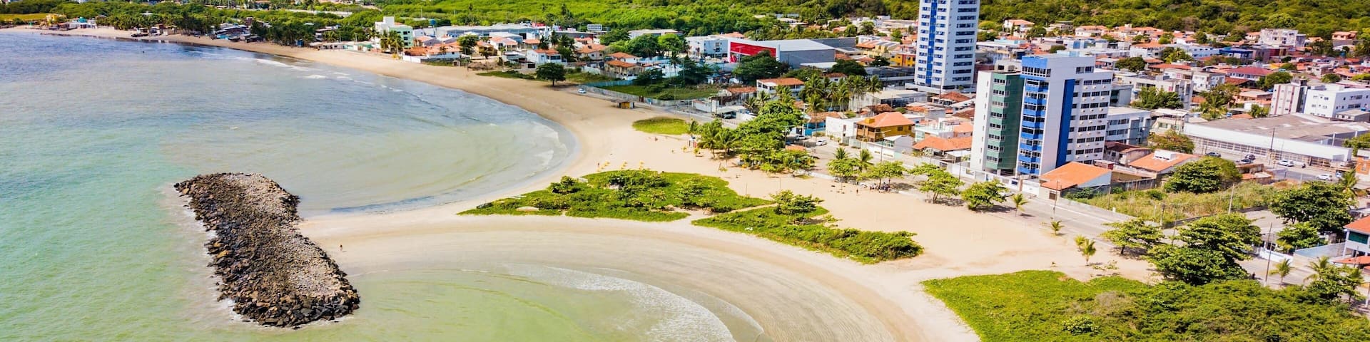 A coastal cityscape with a sandy beach, a rocky outcrop, and a dense urban area with buildings.