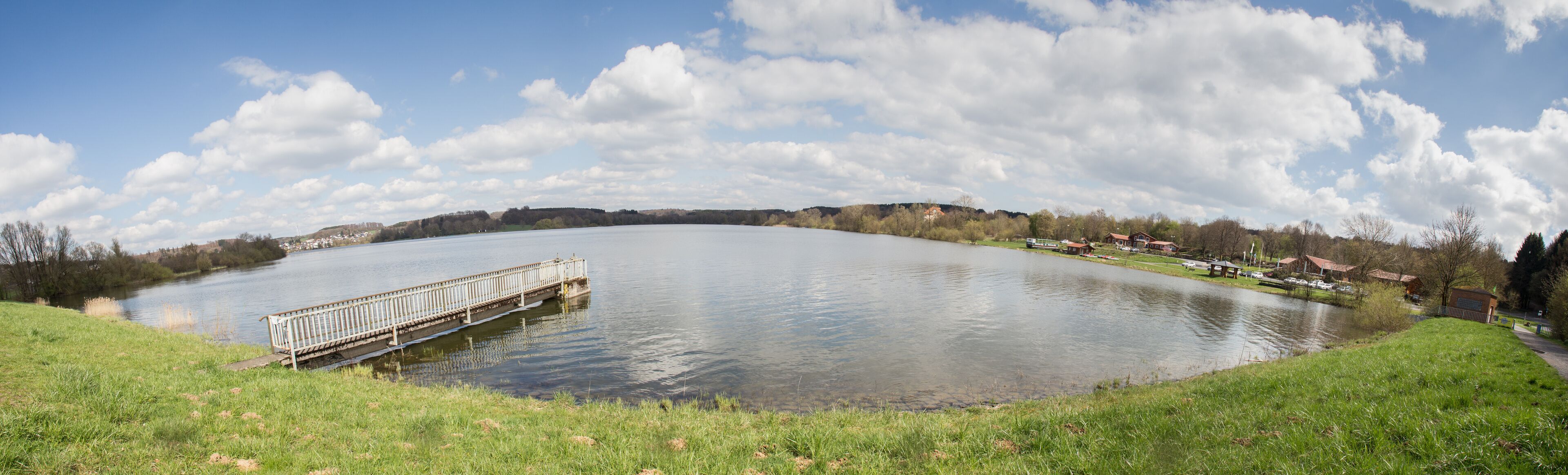 A calm lake with a wooden pier, surrounded by grassy banks and a small village in the distance.
