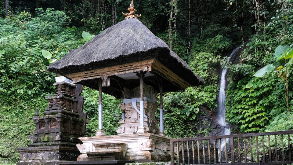 A stone gazebo with a thatched roof, surrounded by lush greenery and a waterfall.