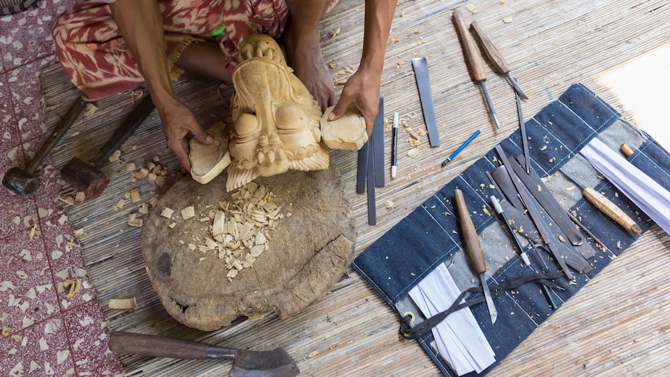 A person carving a wooden statue on a workbench with tools and wood shavings.