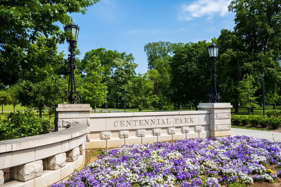 A stone sign reading "CENTENNIAL PARK" surrounded by a flower bed with purple and white flowers.
