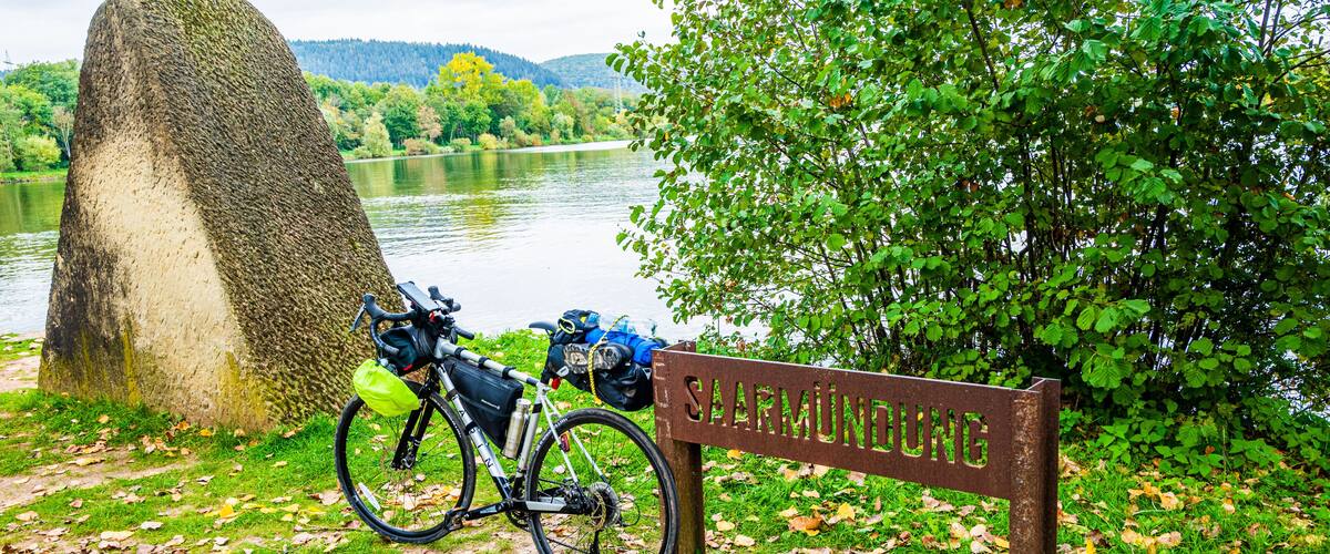 A bicycle parked near a sign that reads 'SAARMÜNDUNG' by a lake with a large rock and trees.