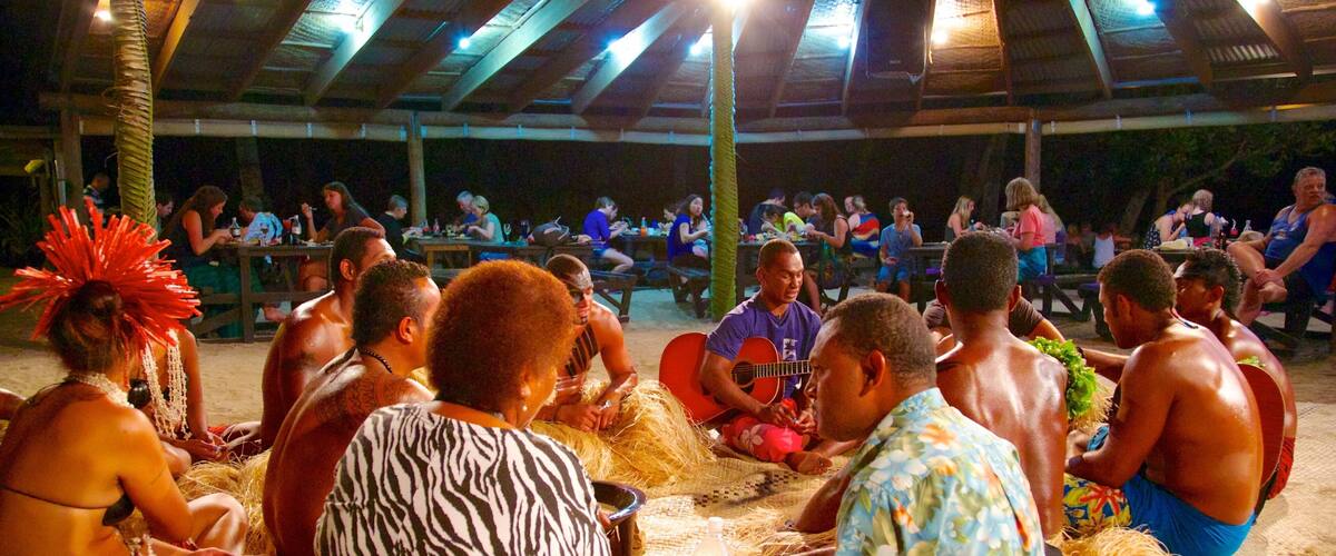 A group of people sitting on hay bales under a thatched roof structure, possibly at a cultural or community event.