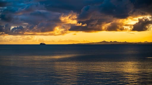 A ship on a calm sea at sunset with a mountain range in the background.