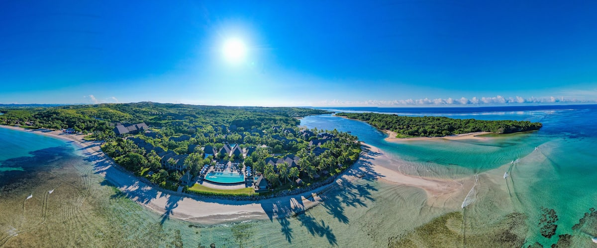 An aerial view of a tropical resort with a swimming pool, palm trees, and clear blue waters.