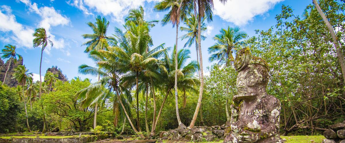 A stone statue in a lush, tropical setting with palm trees and a clear sky.