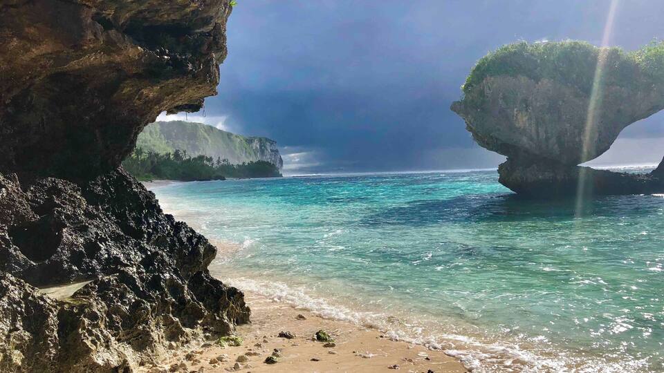 A beach with clear turquoise water, a large rock formation, and a cave entrance.
