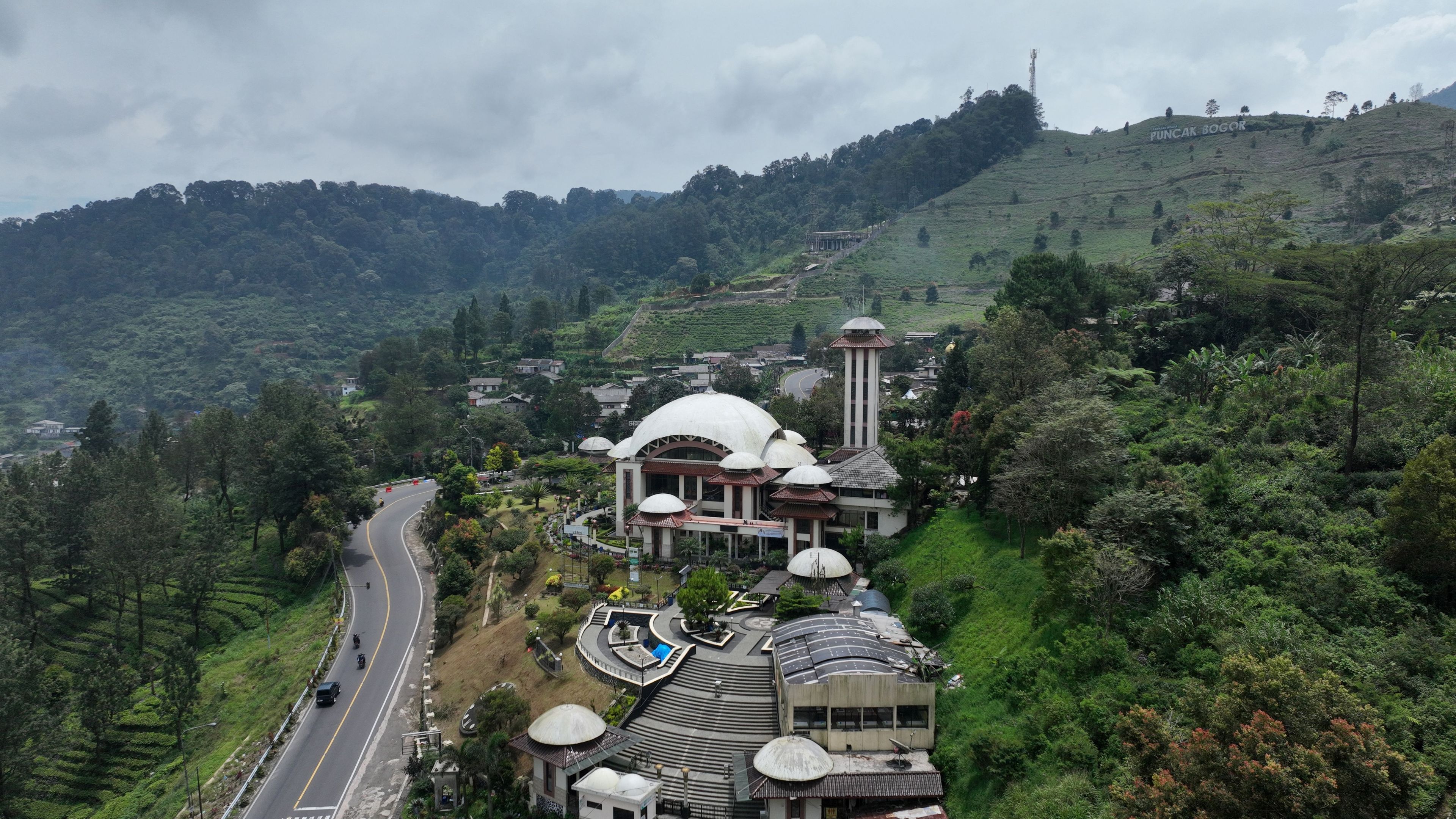 Aerial view of Atta'awun Puncak Mosque surrounded by lush green tea plantations and winding roads in Kecamatan Cisarua, Jawa Barat, Indonesia.