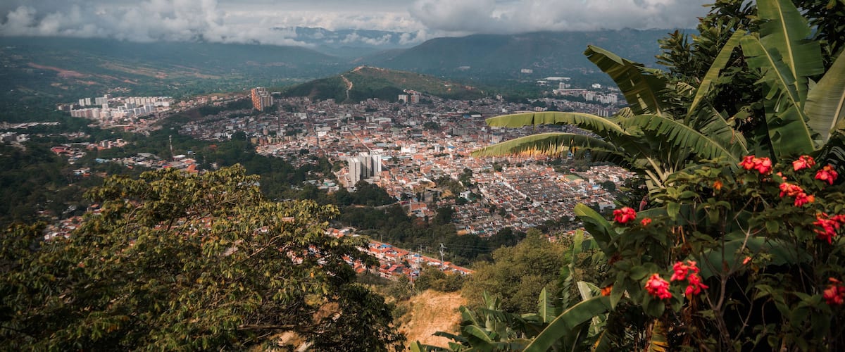 A panoramic view of a city with a prominent cathedral, lush greenery, and distant mountains under a partly cloudy sky.