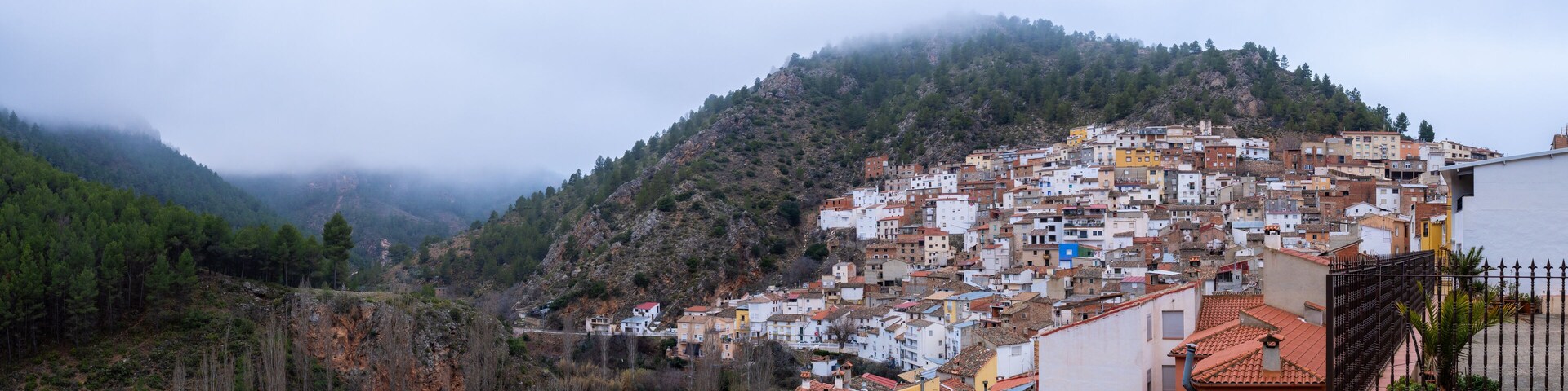 Un pueblo en la ladera de una colina con edificios coloridos, una montaña al fondo y una zona boscosa.