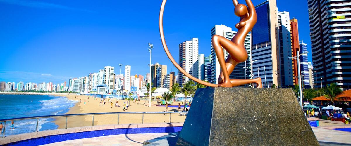 A bronze statue of a figure performing an acrobatic move with a hoop, set against a backdrop of a beach and a city skyline with tall buildings.