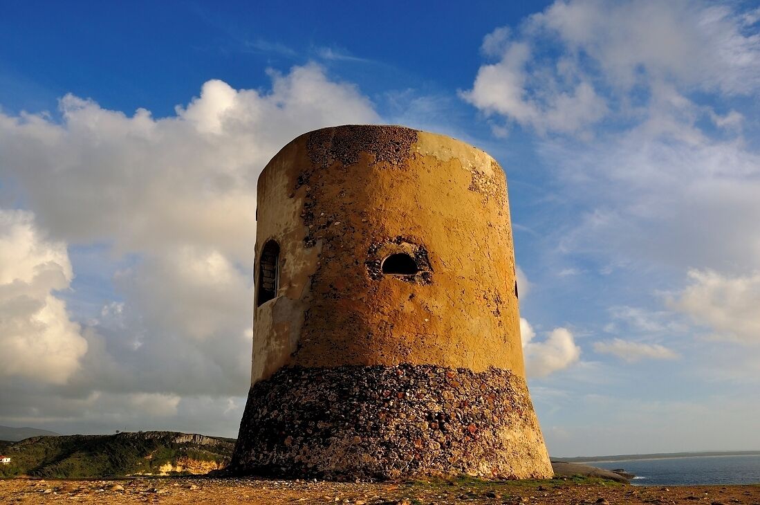 Una torre desgastada con una abertura circular, frente a un paisaje costero con cielo despejado.