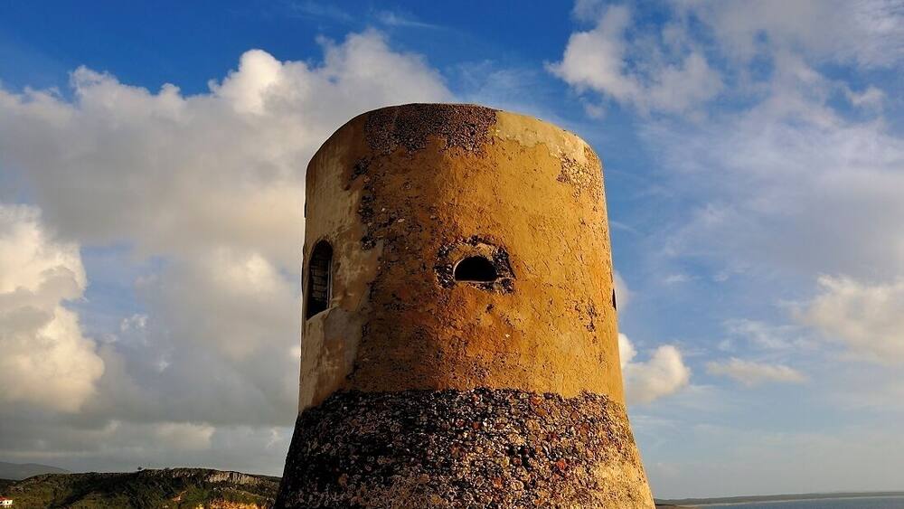 A weathered tower with a circular opening, set against a coastal landscape with a clear sky.