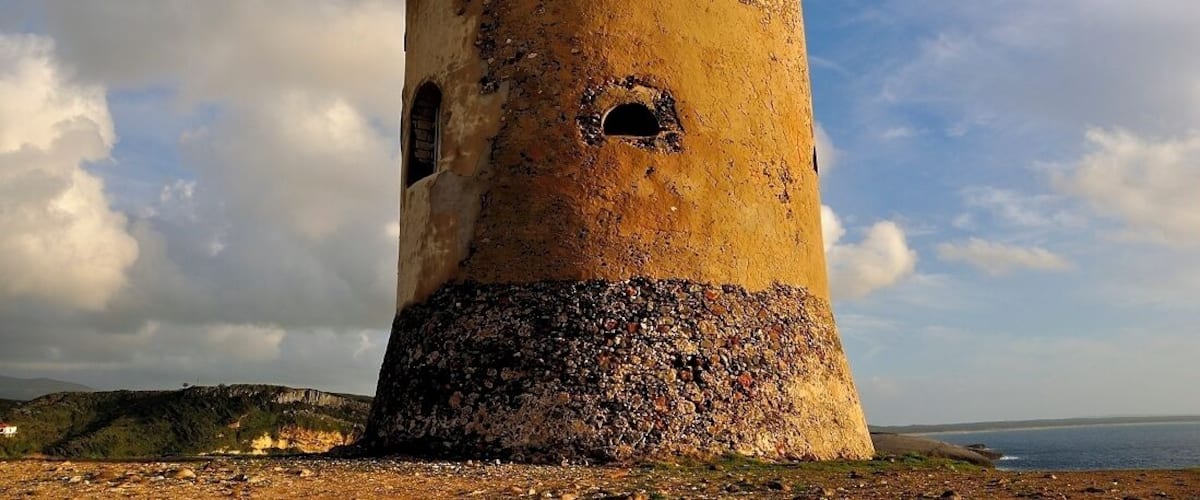 A weathered tower with a circular opening, set against a coastal landscape with a clear sky.