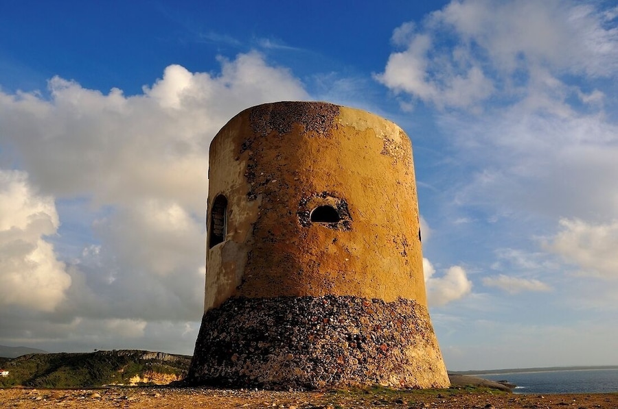 Una torre desgastada con una abertura circular, frente a un paisaje costero con cielo despejado.