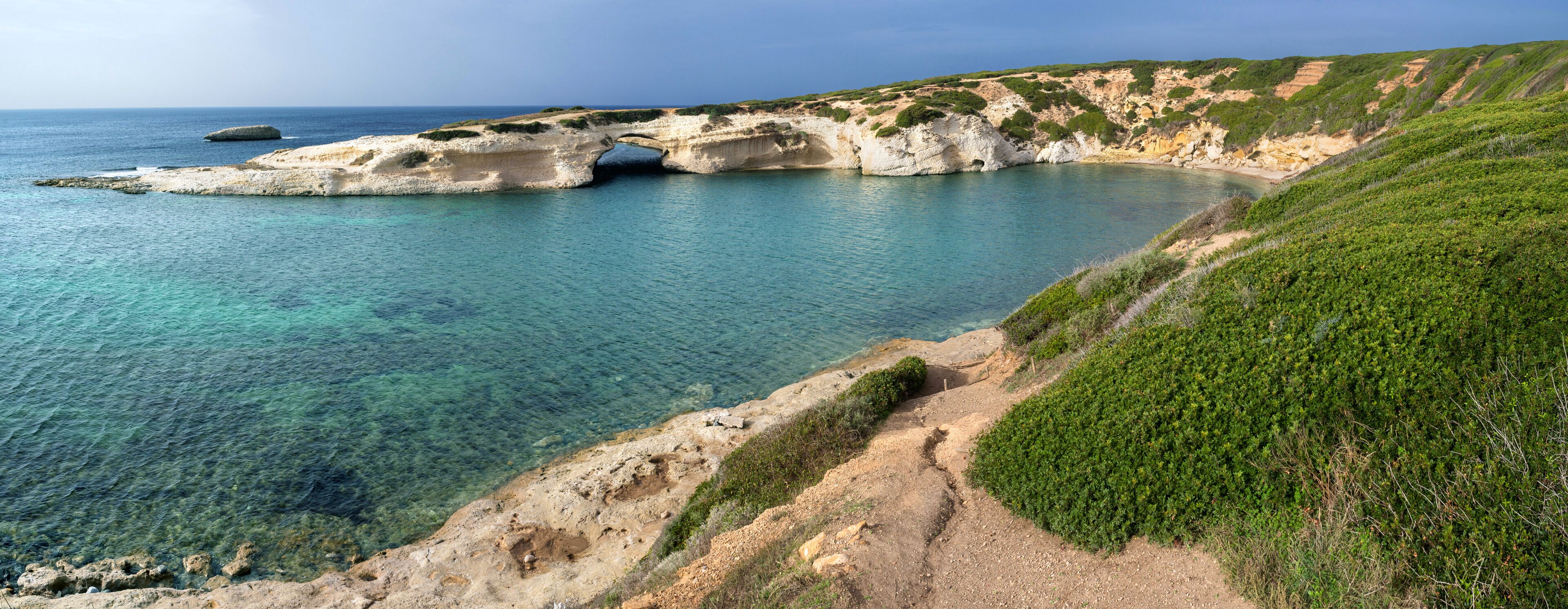 A coastal landscape with clear turquoise waters, rocky cliffs, and a narrow path leading to a white rock formation.