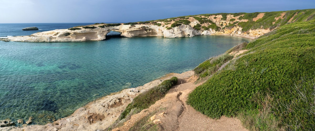 A coastal landscape with clear turquoise waters, rocky cliffs, and a narrow path leading to a white rock formation.
