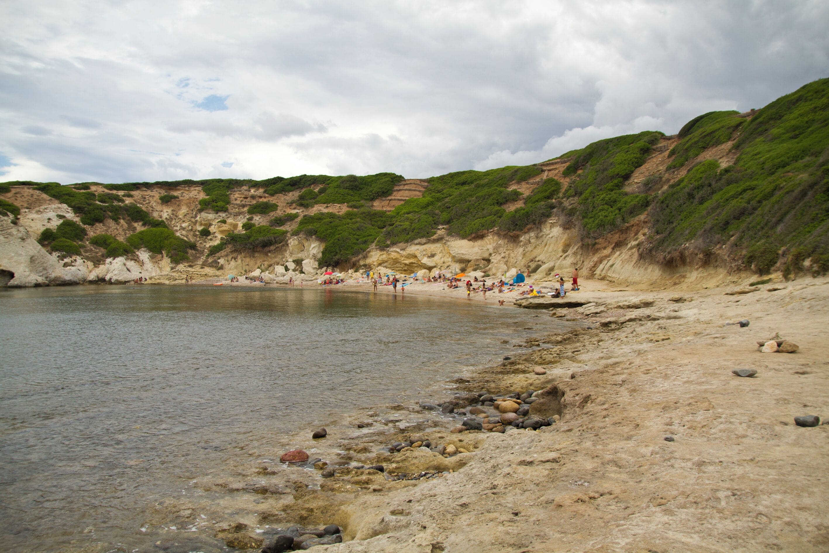 Una playa rocosa con aguas cristalinas, vegetación verde en los acantilados y gente tomando el sol.