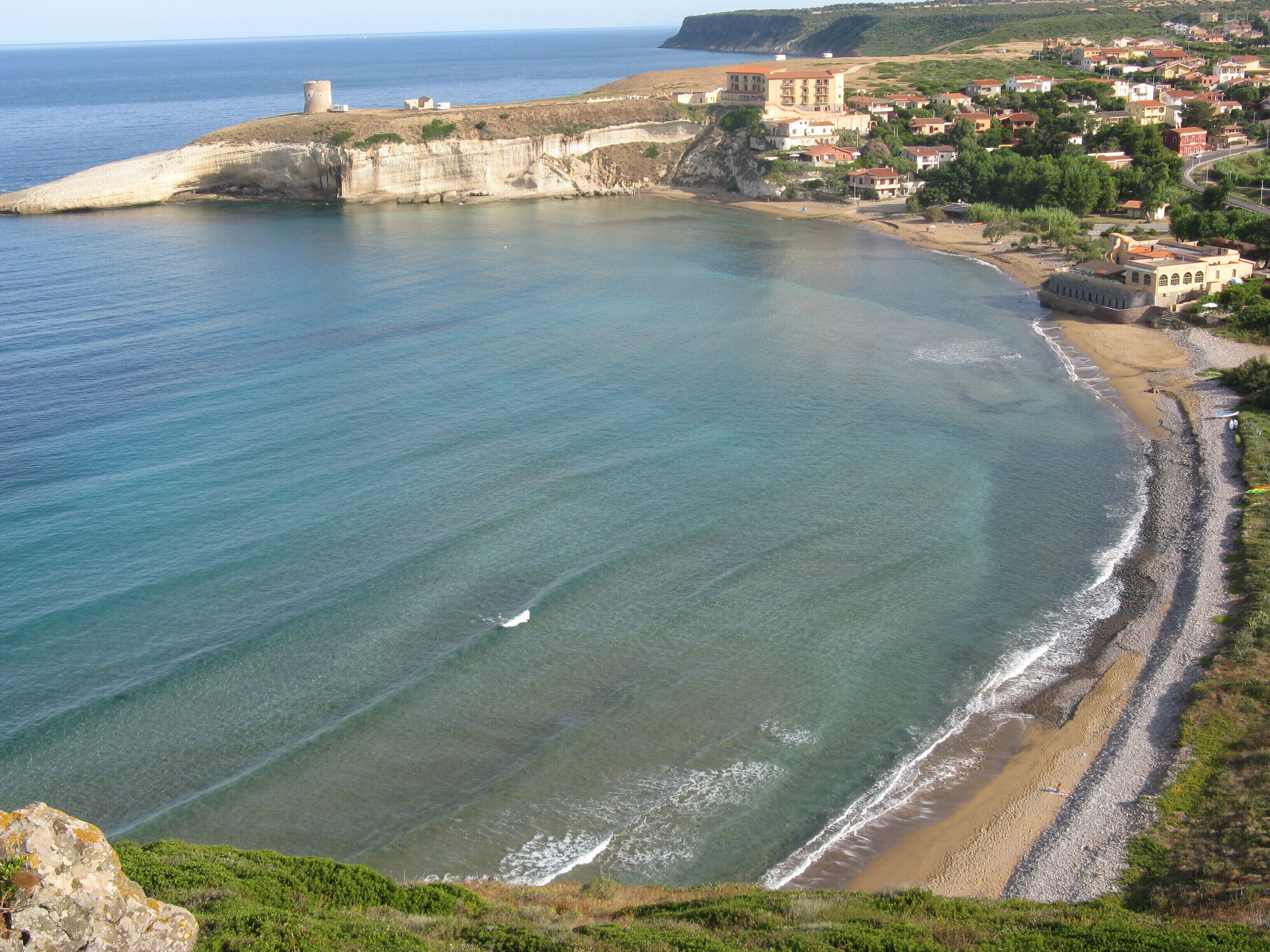 Un pueblo costero con una playa despejada, una estructura fortificada y colinas al fondo.