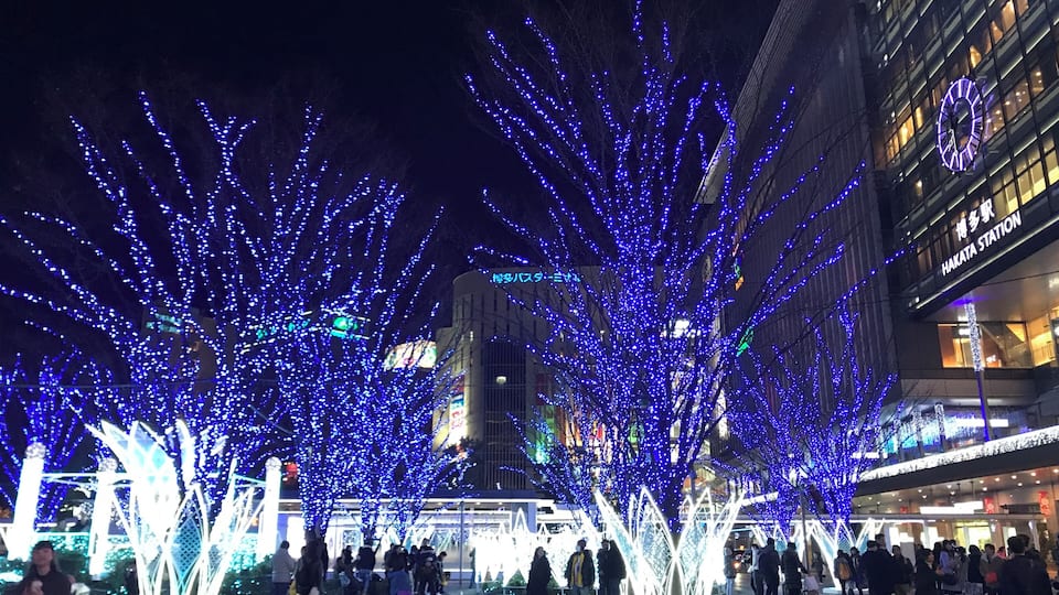 Scena notturna con alberi illuminati e un edificio con la scritta "HAKATA STATION".