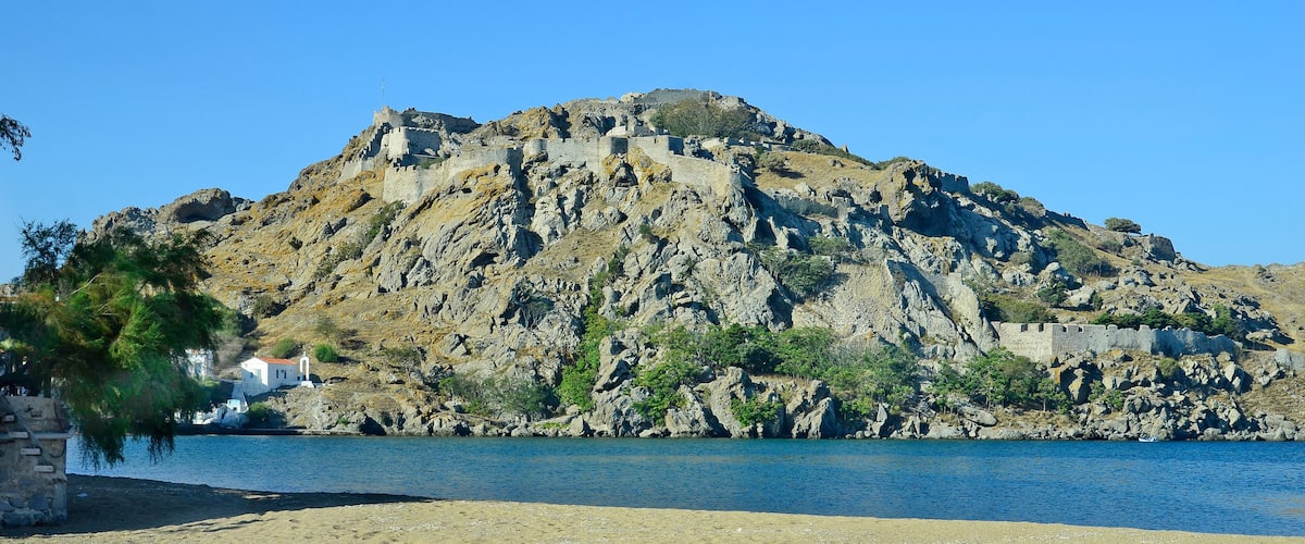 A rocky hill with a castle, a beach, and a calm body of water reflecting the scene.