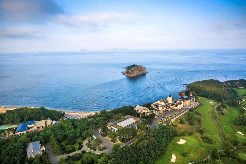 An aerial view of a coastal area with a large estate, a golf course, and a small island in the sea.