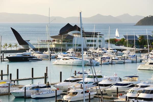 Un port de plaisance où sont amarrés de nombreux yachts, un grand voilier à la voile distinctive et, au fond, un bâtiment à l’architecture moderne.