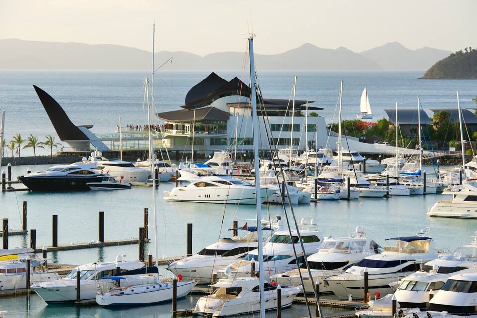 A marina with numerous yachts docked, a large sailboat with a distinctive sail, and a building with a modern design in the background.