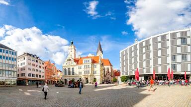 Una plaza adoquinada con edificios históricos, un campanario de iglesia y una construcción moderna.