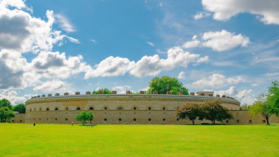 A historic building with a circular design, surrounded by a green field and trees.