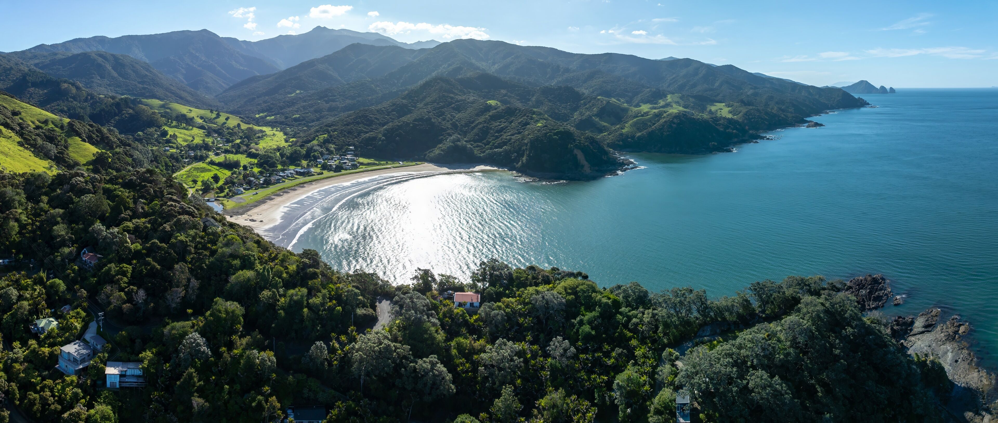 Aerial view of a secluded bay with a sandy beach nestled amongst lush green hills and mountains. Coastal homes are visible along the shoreline. PORT CHARLES, COROMANDEL PENINSULA, NEW ZEALAND