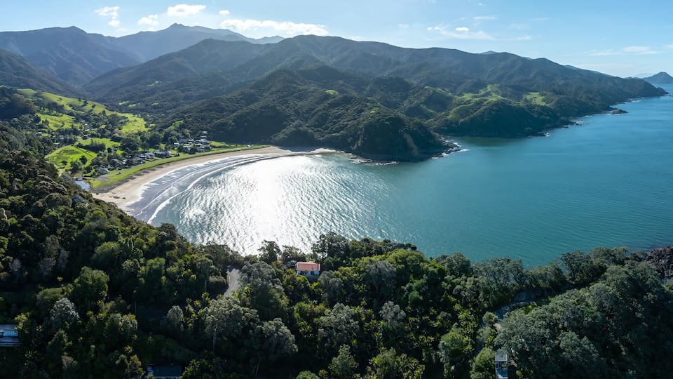 Aerial view of a secluded bay with a sandy beach nestled amongst lush green hills and mountains. Coastal homes are visible along the shoreline. PORT CHARLES, COROMANDEL PENINSULA, NEW ZEALAND