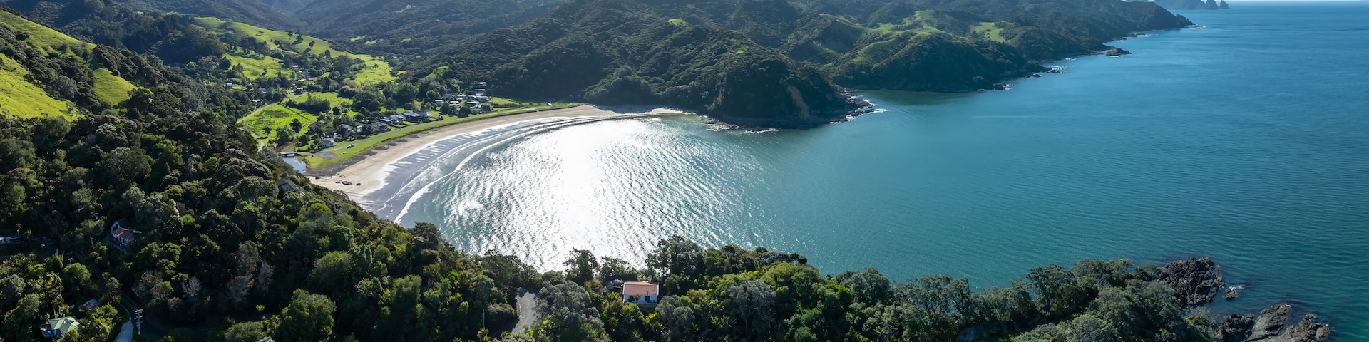 Aerial view of a secluded bay with a sandy beach nestled amongst lush green hills and mountains. Coastal homes are visible along the shoreline. PORT CHARLES, COROMANDEL PENINSULA, NEW ZEALAND