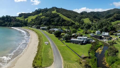 Aerial view of a coastal town nestled in a lush valley. Houses line a road by the beach, showcasing a peaceful seaside community. , PORT CHARLES, COROMANDEL PENINSULA, NEW ZEALAND
