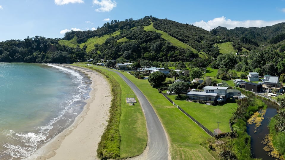 Aerial view of a coastal town nestled in a lush valley. Houses line a road by the beach, showcasing a peaceful seaside community. , PORT CHARLES, COROMANDEL PENINSULA, NEW ZEALAND