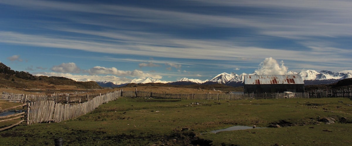 A rural landscape with a wooden fence, a small building, and snow-capped mountains in the background.