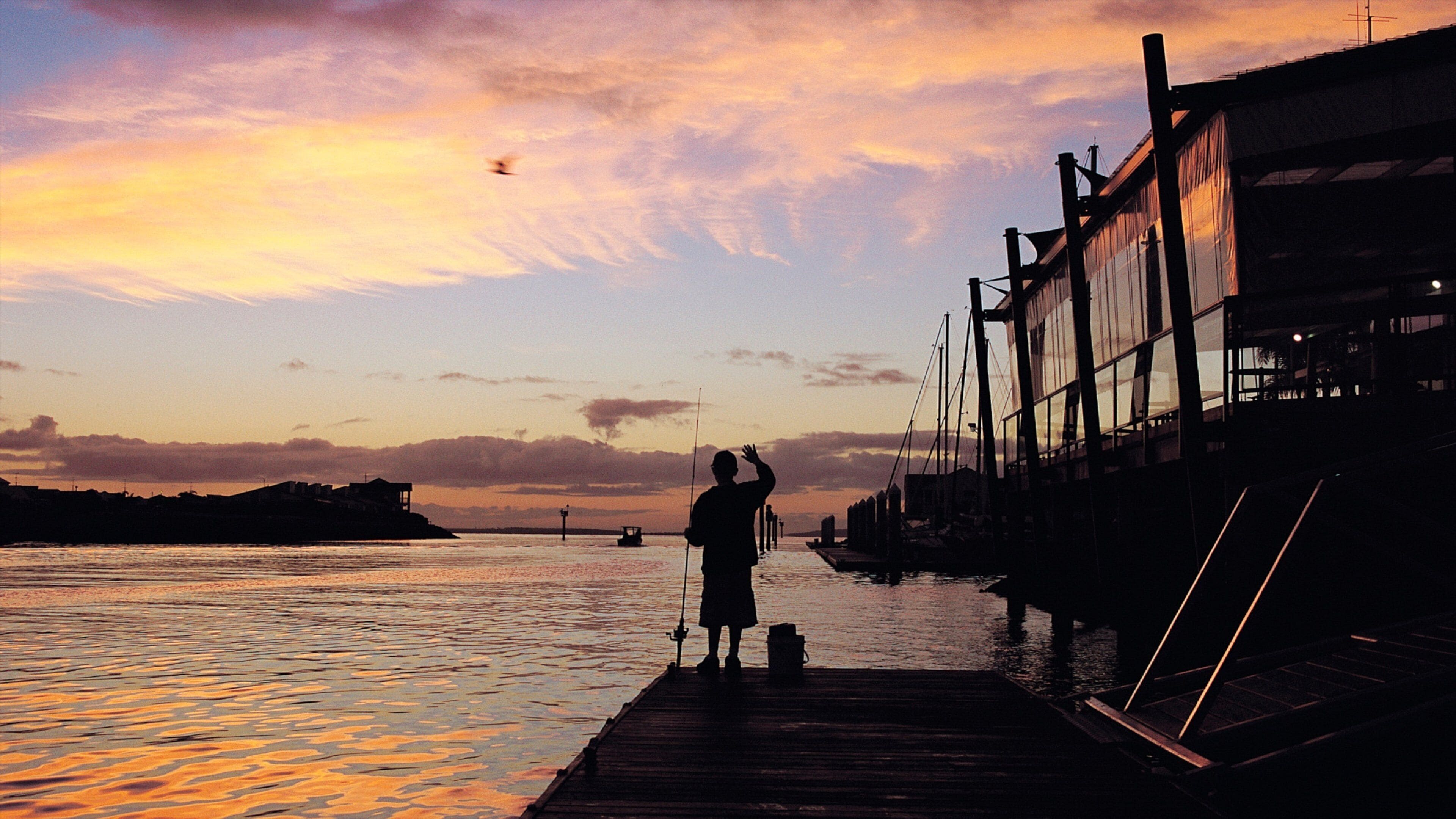 A silhouette of a person on a dock at sunset with a boat in the water and buildings in the background.