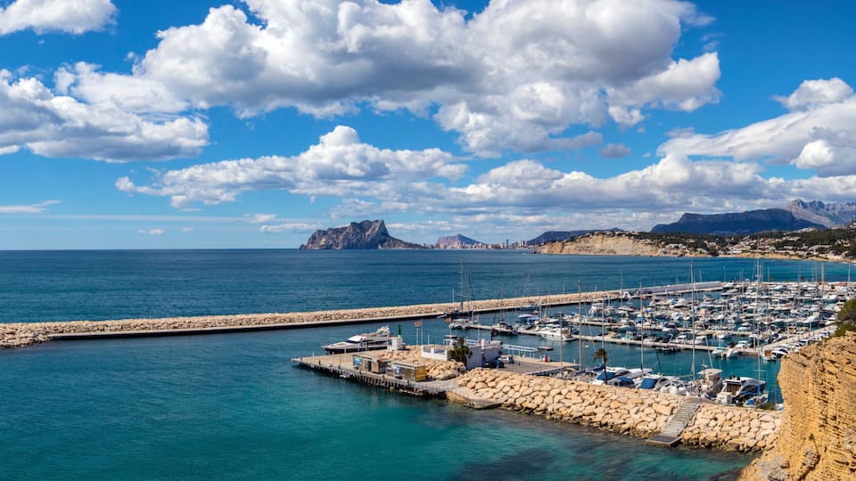 Un puerto costero con barcos, un muelle rocoso y montañas al fondo.