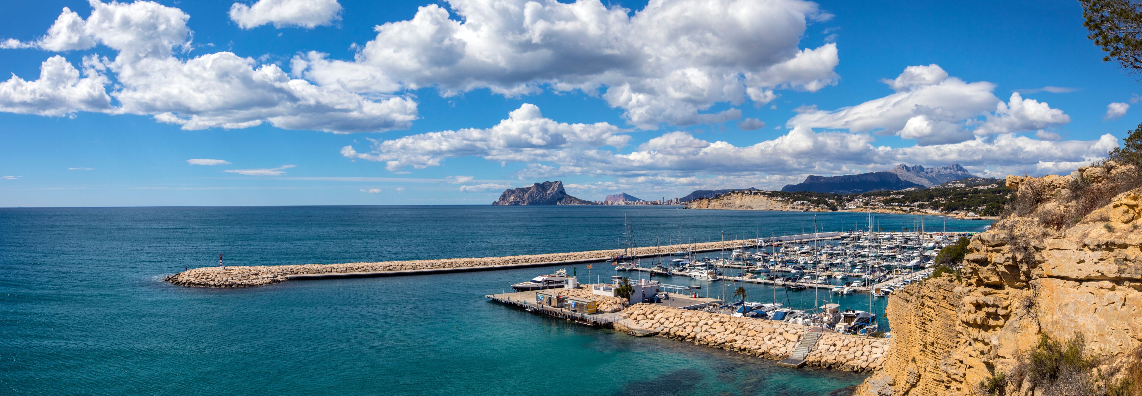 A coastal marina with boats, a rocky pier, and mountains in the background.