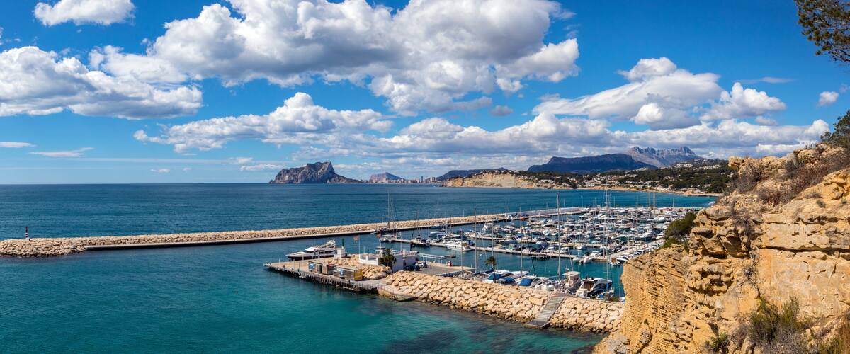 A coastal marina with boats, a rocky pier, and mountains in the background.