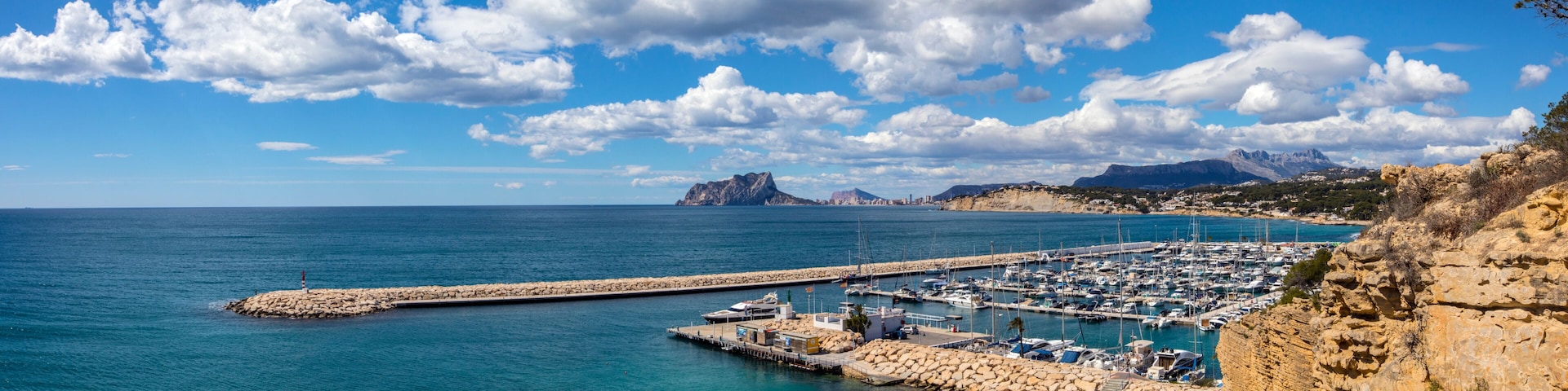 A coastal marina with boats, a rocky pier, and mountains in the background.