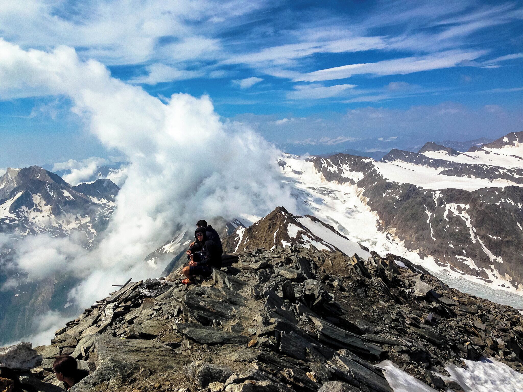 Una persona sentada en la cima de una montaña rocosa, con laderas nevadas y un cielo nublado al fondo.
