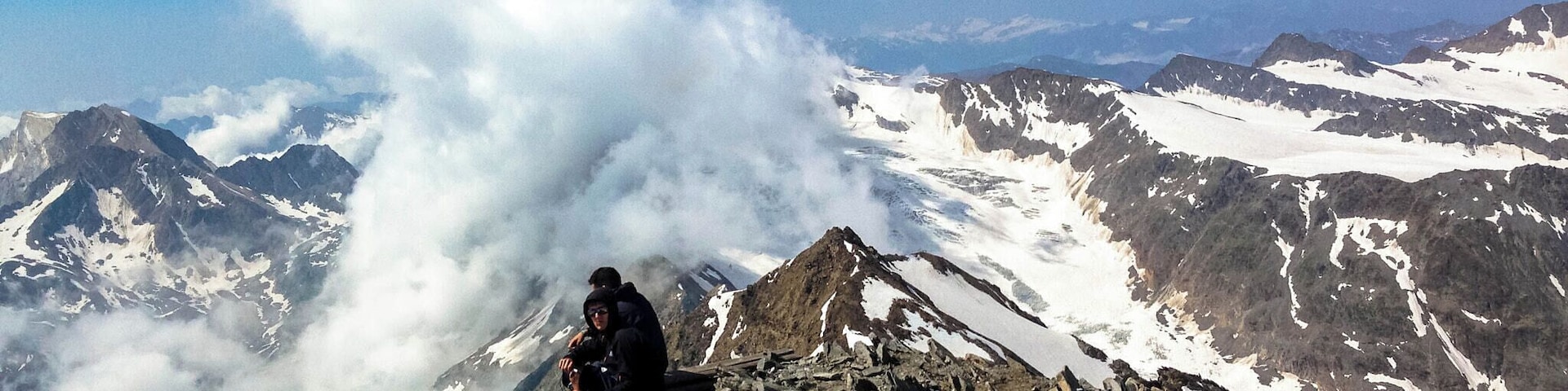 Una persona sentada en la cima de una montaña rocosa, con laderas nevadas y un cielo nublado al fondo.